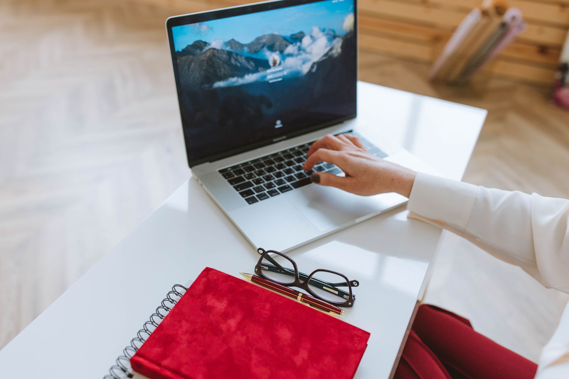 Une personne travaille sur un ordinateur portable avec un carnet rouge et des lunettes sur une table blanche.