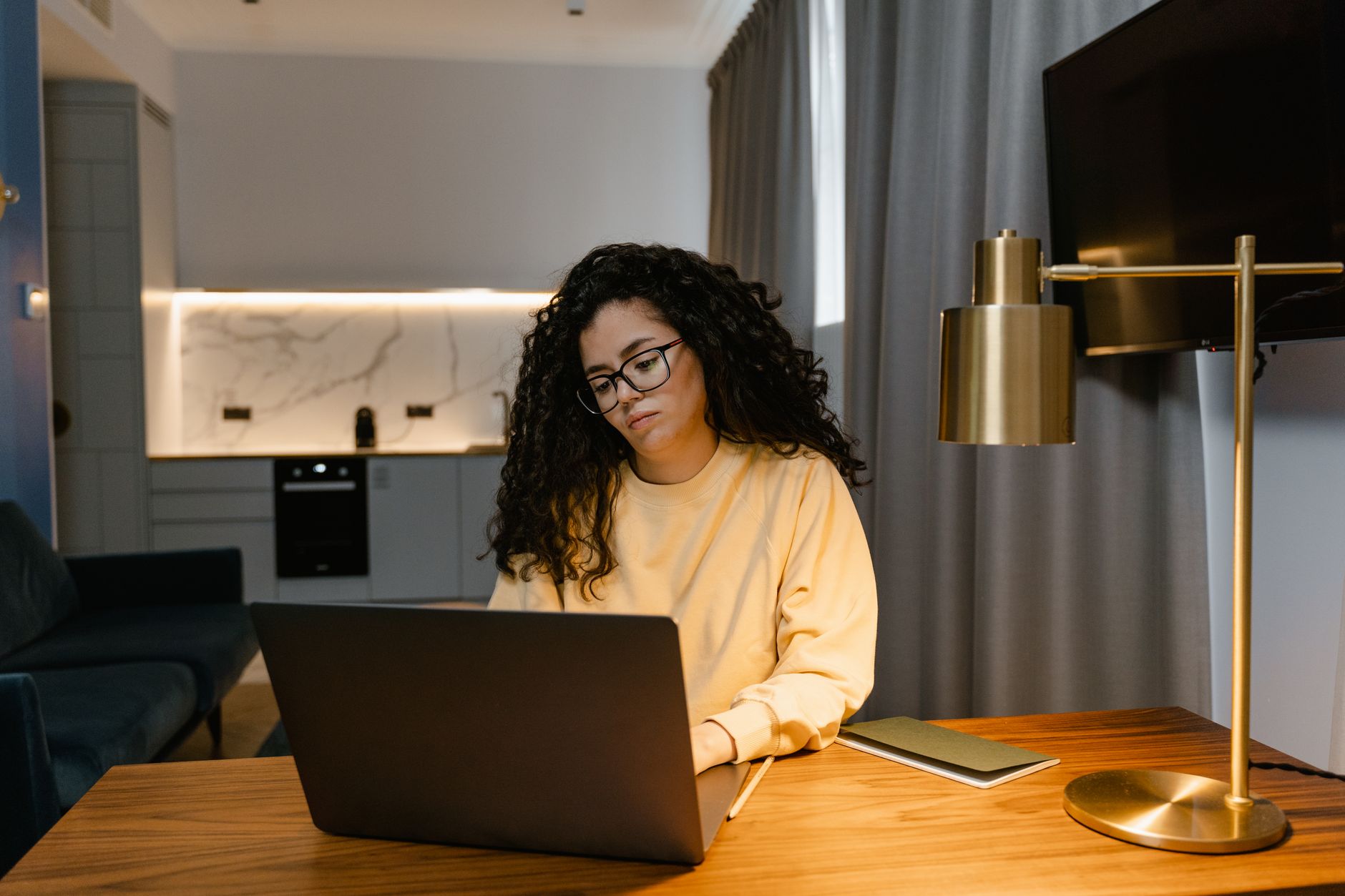 Femme en sweat-shirt jaune utilisant un ordinateur portable à la maison, illustrant le travail à distance dans un intérieur cosy et moderne.